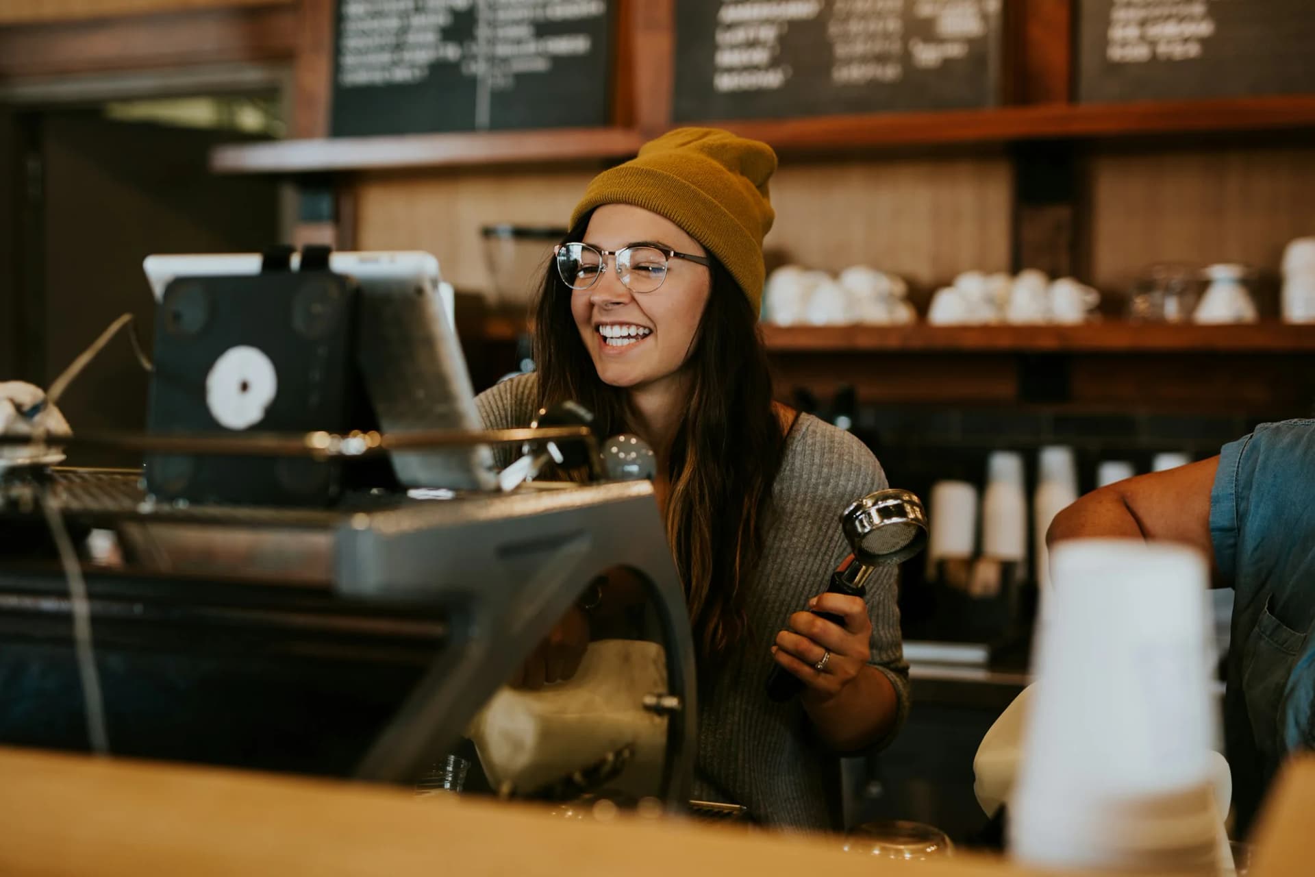 Nevető fiatal barista lány mustársárga sapkában, kávéfőzővel a kezében egy kávézó pultja mögött